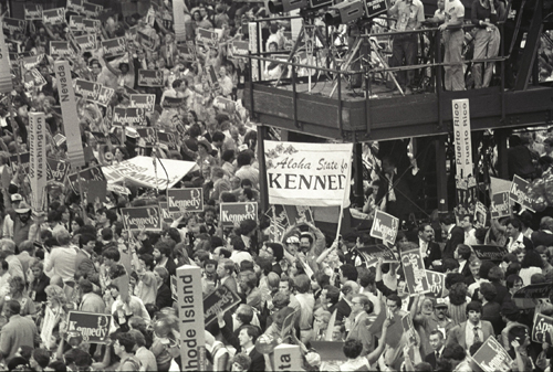 A wide shot above the crowd reveals a sea of Kennedy signs