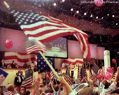 an american flag waves across the foreground and the Republican podium in in the background as balloons and confetti drop from the rafters