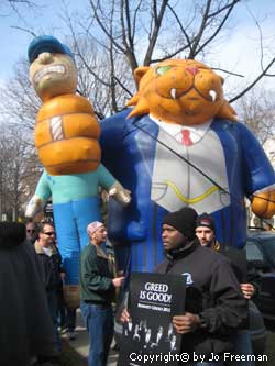 a large inflatable fat cat wears a businesss suit and chokes a worker in one hand, while a nearby person holds a sign reading Greed is Good with a picture of Romney