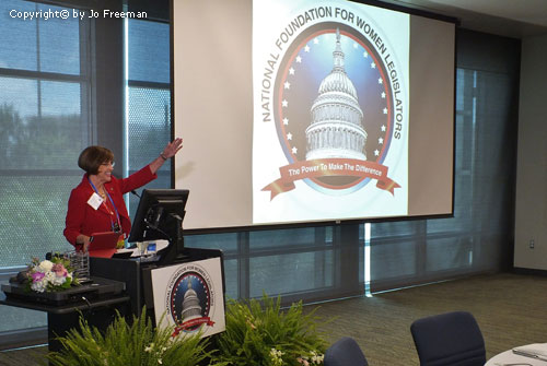 A Woman stands at a podium and gestures toward a screen