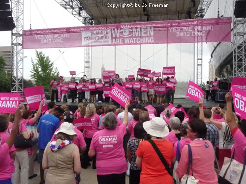 many people wearing pink shirts stand on or in front of a stage with the mottos Yes We Plan and We Are Voting Obama