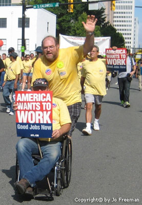 a man in a parade waves and pushes another man in a wheelchair who holds a sign reading America Wants to Work Good Jobs Now