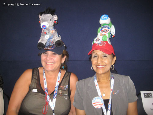 Two ladies wear festive plush toys on baseball hats with political buttons pinned to them