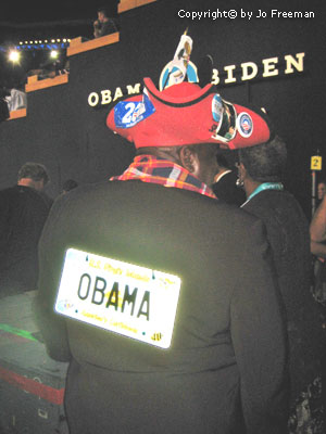 A man wearing a red trefoil hat displays a liscence plate on his back that reads OBAMA