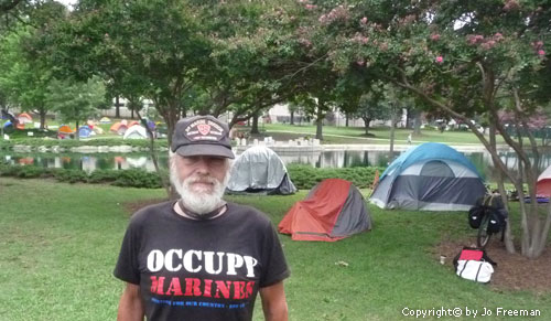 a man stands in front of tents in a park setting near a pond