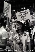 A female deligate wears an ERA sash and shouts while holding a sign, many others hold thesame sign in the crowd