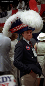 Man wears gigantic and flamboyent three pointed hat reading Kennedy