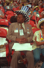 African-American male deligate wears a striped and polkadotted blue and white top hat with Kennedy written on an arch above it