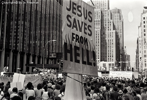 a sign in the foreground reads Jesus saves from hell, in the background are many protestors, facing away form the sign and towards a large stage