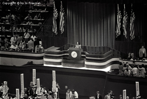 An expansive shot shows a small podium with a person behind it on a vast stage flanked by american flags.