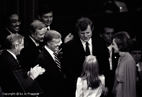 Kennedy and Carter stand next to each other and smile while nearby people clap.  The two politicians appear to be speaking with someone's daughter.