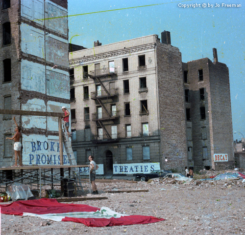 condemned buildings have painted on signs.  one reads Broken Promises another reads Broken Treaties, a third building simply reads Decay.  A stage is beting set up in the foreground.