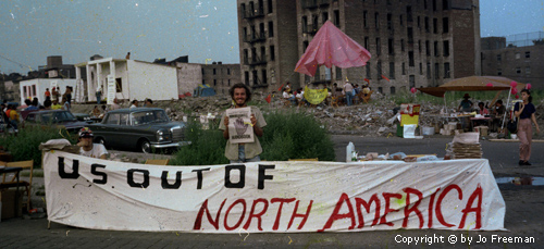 with the condemned high-rises and mountains of rubble in the backgorund, a young man stands in front of a huge sign in the foreground which reads US out of North America