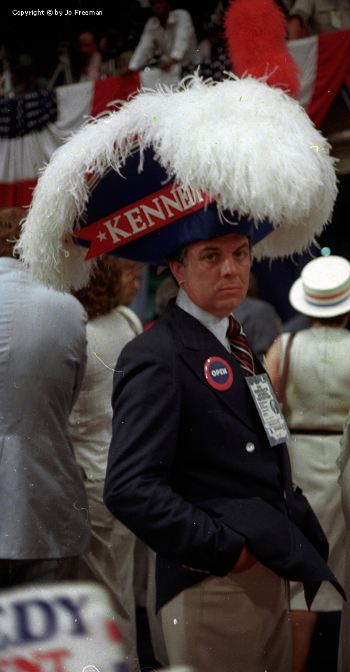 A male deligate wears a gigantic blue and feathery three cornered hat reading Kennedy.