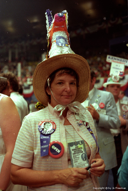 A female deligate wears a large donkey pinata on top of her hat.  the donkey has a photo of Kennedy