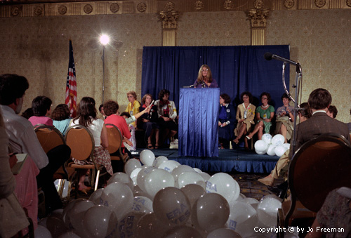 Gloria Steinem at the podiium in front of a full audence and many balloons in the aisle