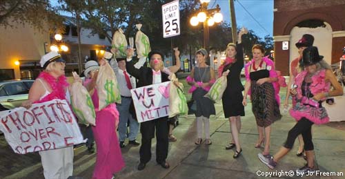 People finely dressed with pink accent pieces hold moneybags and signs ironically supporting Romney's profits over people