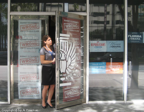 a woman stands in the doorway of a storefront covered with anti-Romney, pro-Obama posters