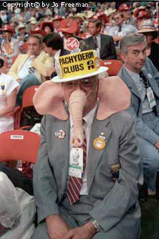 a man wears a prosthetic elephant trunk and elephant ears and a hat reading Pachyderm Clubs