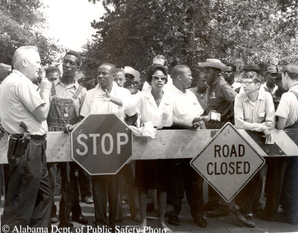 a primarily african american crowd stands behind barracades, in the foreground is a policeman